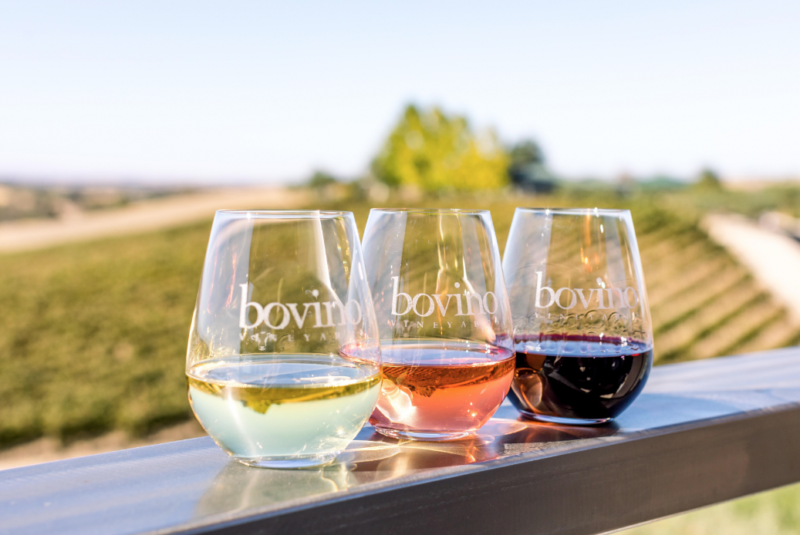 Three wine glasses (from left to right: a white wine, rose wine, and a red wine) sitting on a wooden ledge overlooking a vineyard