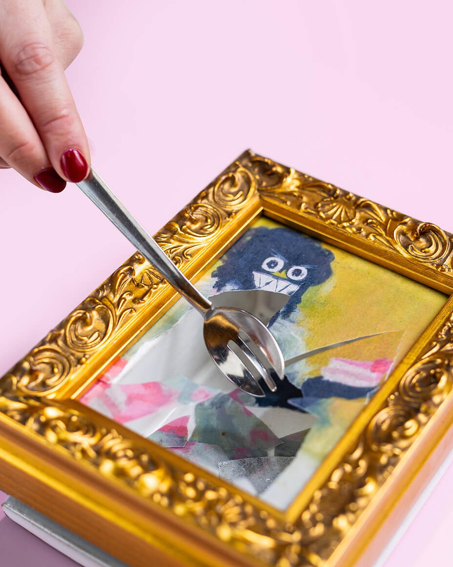 Hand with spoon breaking the surface of a plated dish that looks like a picture frame with colorful art