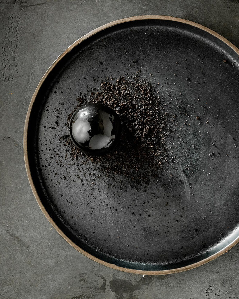 Black spherical plated dish with charcoal-colored crumbs on a plate and counter of the same colors
