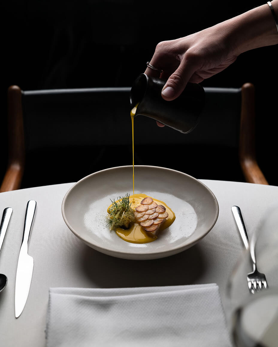 Hand pouring small carafe of yellow sauce onto plate with turbot dish