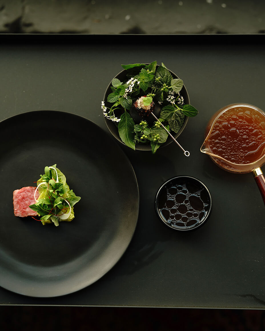 Four dishes containing wagyu, broth, herbs and skewered meat, and sauce on a stark black background