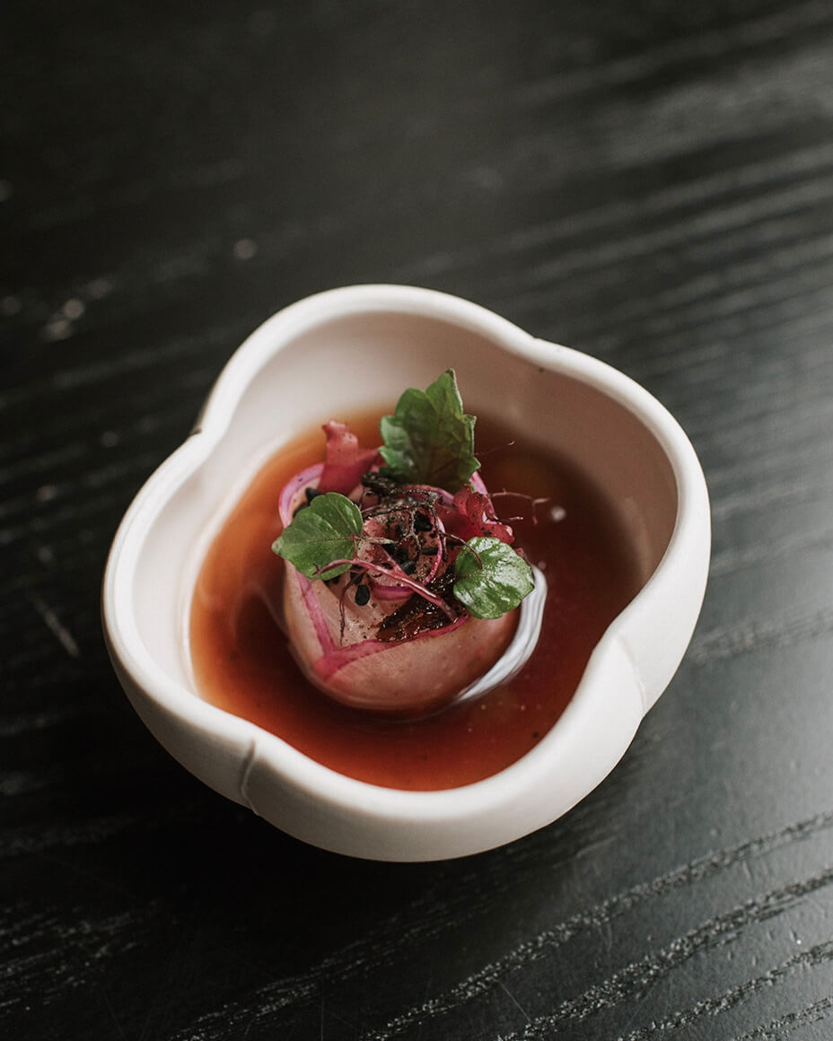 Small four petal flower-like bowl containing hiramasa fish plated like a rose in broth