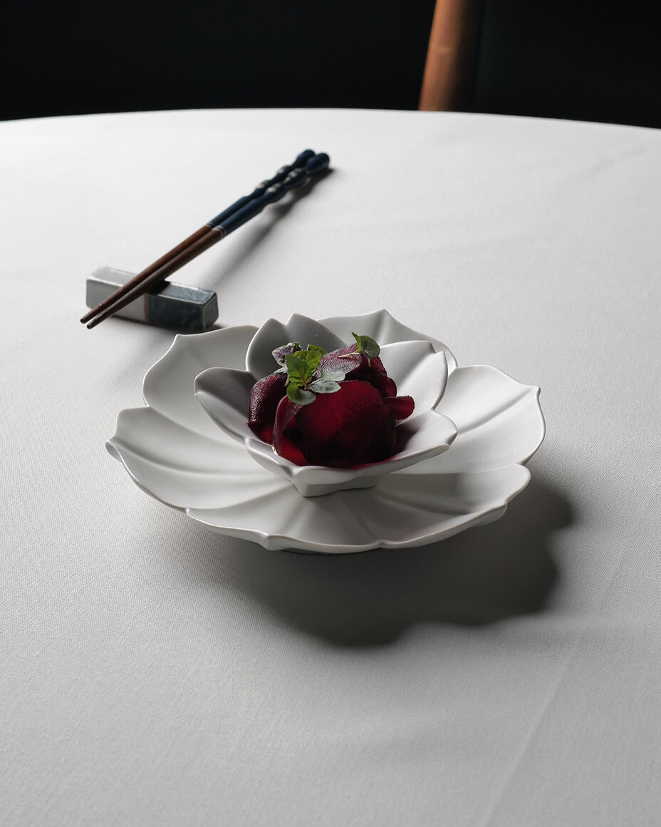 Beet dish plated on a two-tier white flower plate next to a set of chopsticks on a white linen table