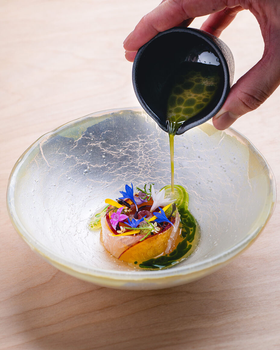 Hand pouring green oily sauce onto colorful plated dish on a pearlescent white bowl with a crackle pattern