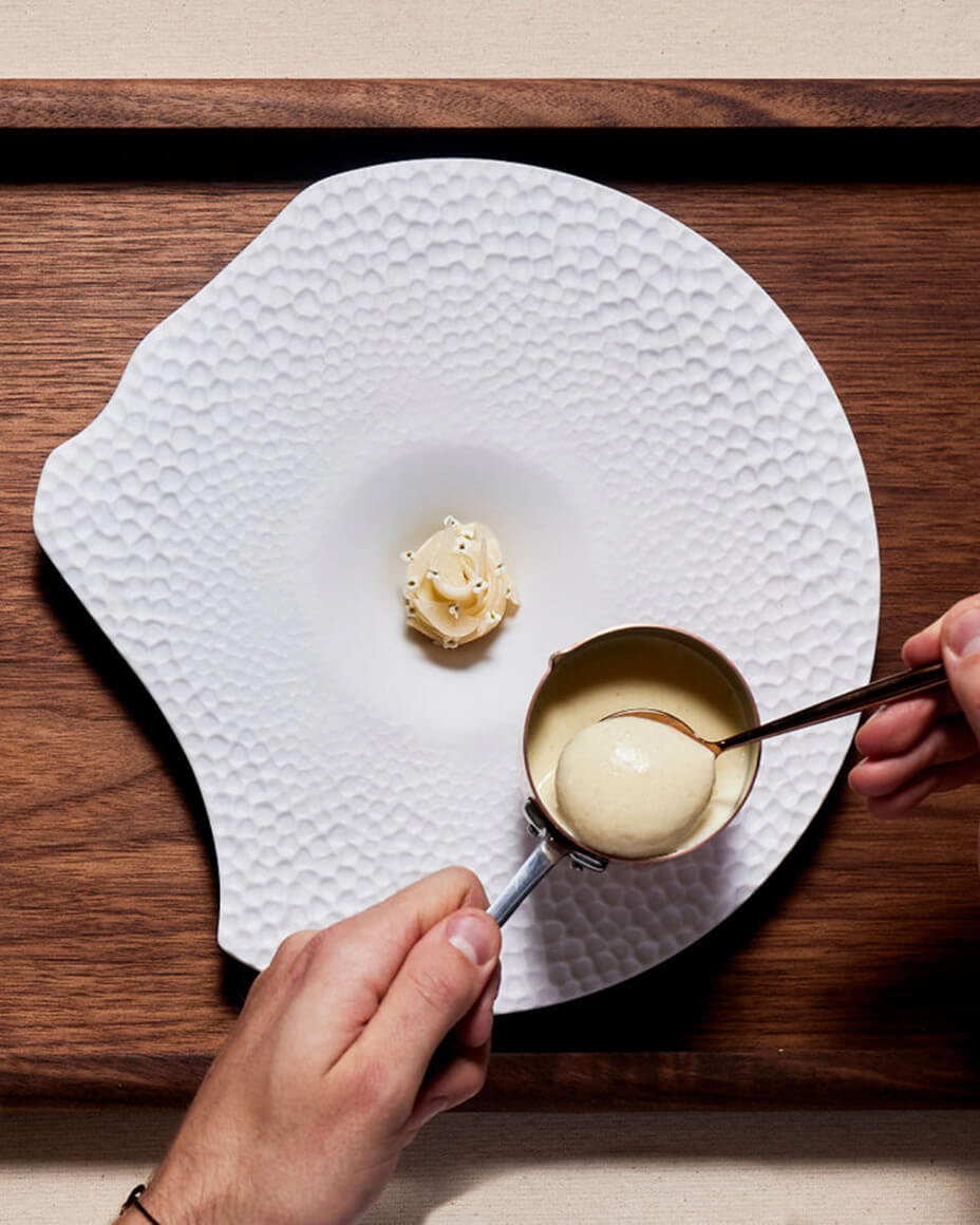 Hands plating a white, ice cream like substance from a mini pot onto a uniquely shaped textured white plate