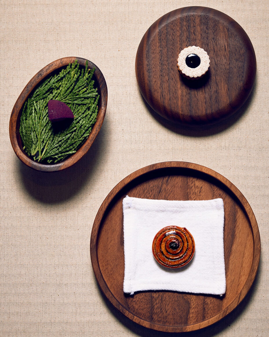 Three wooden plates with singular food items on top, one bite that resembles a beet wedge, another a linzer cookie, and a swirled pastry