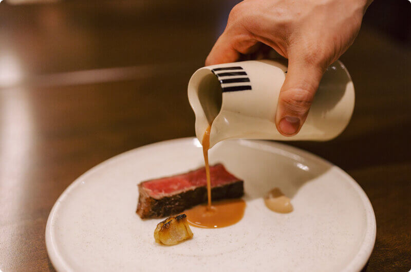 Hand pouring a sauce out of a small carafe onto a plated dish with steak.