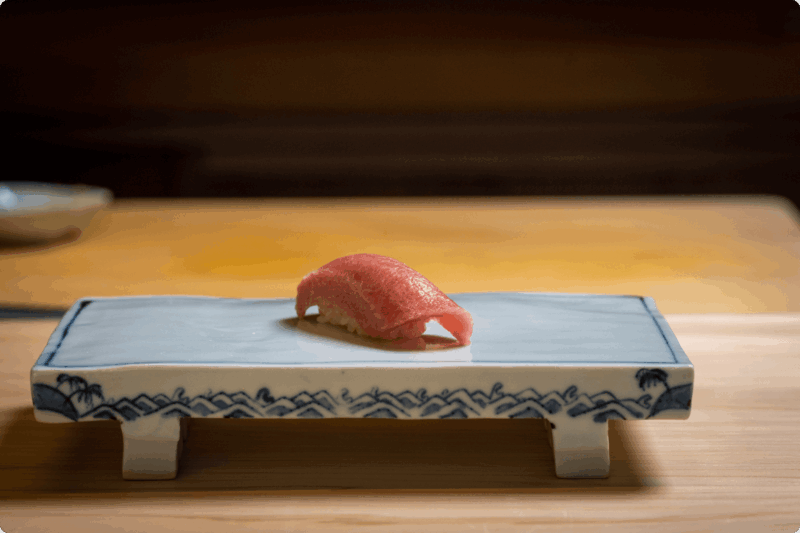 Single piece of tuna nigiri on an ornate rectangular white and blue plate