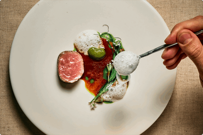 Hand spooning foam onto plate with a slice of meat, red sauce, and herbs