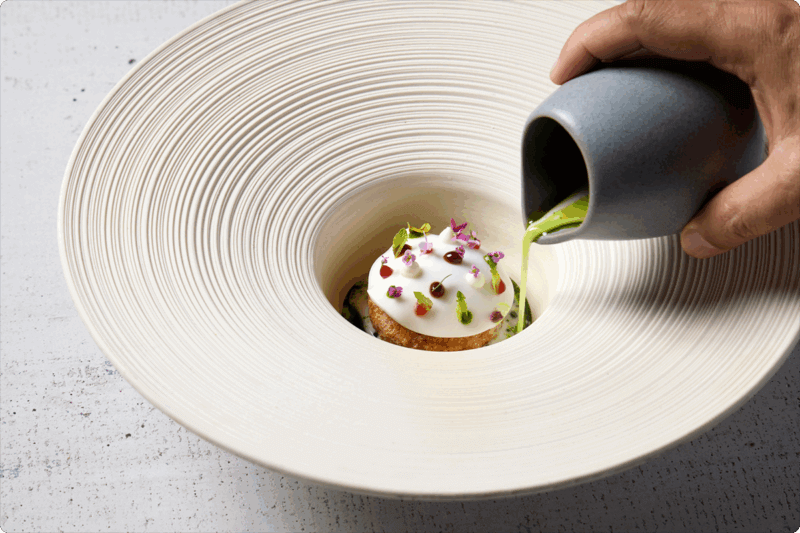 Hand pouring a green liquid into a bowl of a small, beautifully plated dish